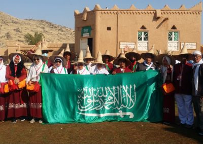 Rombongan jamaah umrah berfoto dengan bendera Arab Saudi di depan Almoogi Museum, mengenakan pakaian seragam merah dan topi tradisional