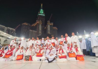 Rombongan jamaah umrah sedang berfoto bersama di area Masjidil Haram, Mekkah, dengan latar belakang Menara Jam dan pencakar langit.