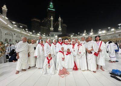 Rombongan jamaah umrah sedang berfoto bersama di area Masjidil Haram, Mekkah, dengan latar belakang Menara Jam dan pencakar langit.